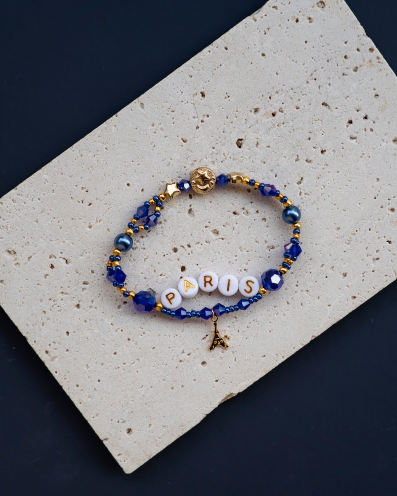 May include: A blue and gold beaded bracelet with a charm of the Eiffel Tower and white beads spelling out "PARIS".