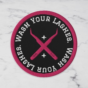 May include: Embroidered patch with a black center and a hot pink border. The text "WASH YOUR LASHES" is in white, surrounding a crossed pair of pink eyelash extension tools and two white stars.