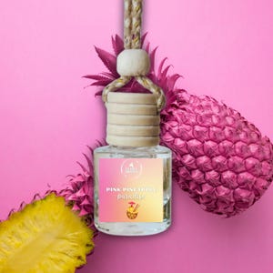 May include: A clear glass car air freshener with a wooden cap and a rope hanger. The label reads "Pink Pineapple Paradise" with a pineapple illustration. The background features a pink pineapple and a pink backdrop.