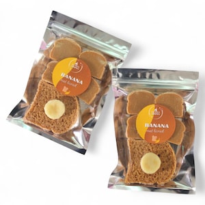May include: Two sealed bags of banana nut bread slices. Each bag contains several slices of bread and a round sticker with the words "Banana nut bread". One slice has a round topping.