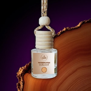 May include: A clear glass car air freshener bottle with a wooden cap and a beige rope for hanging. The label reads "Mahogany Teakwood" with a wood grain graphic. The background is a gradient of purple and brown.