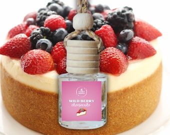 May include: A clear glass air freshener bottle with a wooden cap and a pink label that reads "Wild Berry Cheesecake." The bottle hangs in front of a cheesecake topped with fresh berries. The cheesecake sits on a white plate.