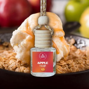 May include: A clear glass air freshener bottle with a wooden cap and a rope hanger. The label reads "APPLE CRISP" with a graphic. The bottle is hanging in front of a dessert with ice cream and caramel.