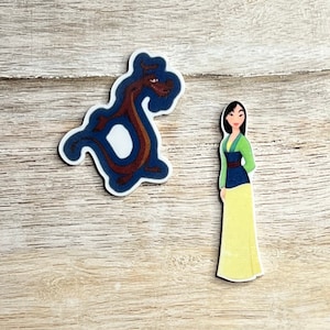 Puede incluir: Una pegatina con una imagen de dibujos animados de Mulan, una joven con el pelo largo y negro, que lleva un vestido verde y azul y una falda amarilla. Está de pie junto a un dragón rojo con alas azules.