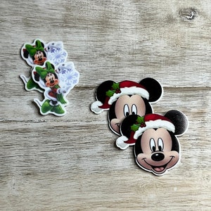 May include: Three Christmas-themed Mickey Mouse stickers. The stickers are round and feature Mickey Mouse wearing a Santa hat. The stickers are on a light brown wooden surface.