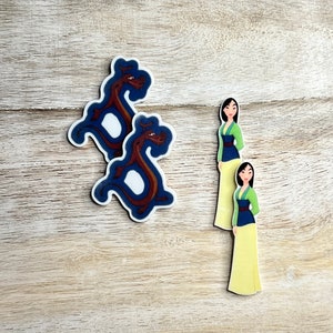 Puede incluir: Dos pegatinas con un dragón rojo y una mujer con un vestido verde y azul. Las pegatinas están sobre una superficie de madera marrón claro.