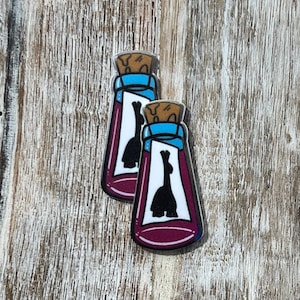 May include: Two bottle-shaped pins with a dark purple liquid and a black llama silhouette inside. The bottles have a blue rim, a cork stopper, and a white label. The pins are set against a light brown wooden background.
