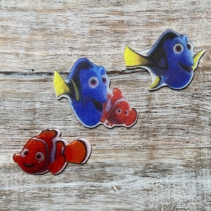 Puede incluir: Tres pegatinas de colores en forma de pez, dos azules y una naranja, con contornos blancos. Los peces azules tienen aletas amarillas y ojos negros. El pez naranja tiene rayas blancas y ojos negros.