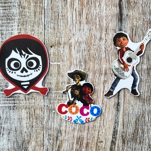 Może przedstawiać: Trzy kolorowe naklejki z postaciami z filmu animowanego Coco. Naklejki przedstawiają Miguela grającego na gitarze, Hectora i Dantego oraz tytułową kartę z napisem "Coco".