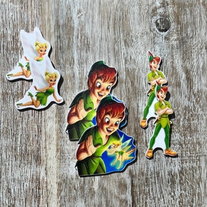 May include: A set of five Peter Pan and Tinkerbell character cutouts. The cutouts are printed on white cardstock and feature Peter Pan in a green tunic and brown pants, Tinkerbell with her wings spread, and a green and brown background.