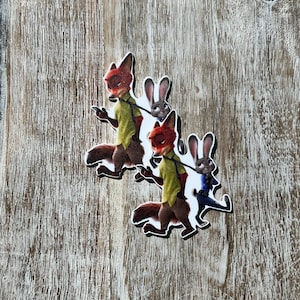 以下が含まれることがあります： 映画ズートピアの2人のアニメキャラクター。緑のシャツを着た赤いキツネと、青いシャツを着た白いウサギ。キャラクターはお互いの尻尾をつかんでいます。