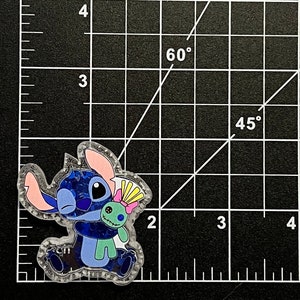 May include: A clear, glittery pin featuring the character Stitch from the animated film Lilo & Stitch. Stitch is hugging a green voodoo doll. The pin has blue, pink, and yellow accents and is set against a black grid background.