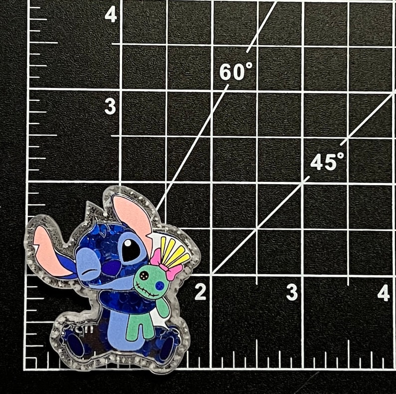 May include: A clear, glittery pin featuring the character Stitch from the animated film Lilo & Stitch. Stitch is hugging a green voodoo doll. The pin has blue, pink, and yellow accents and is set against a black grid background.