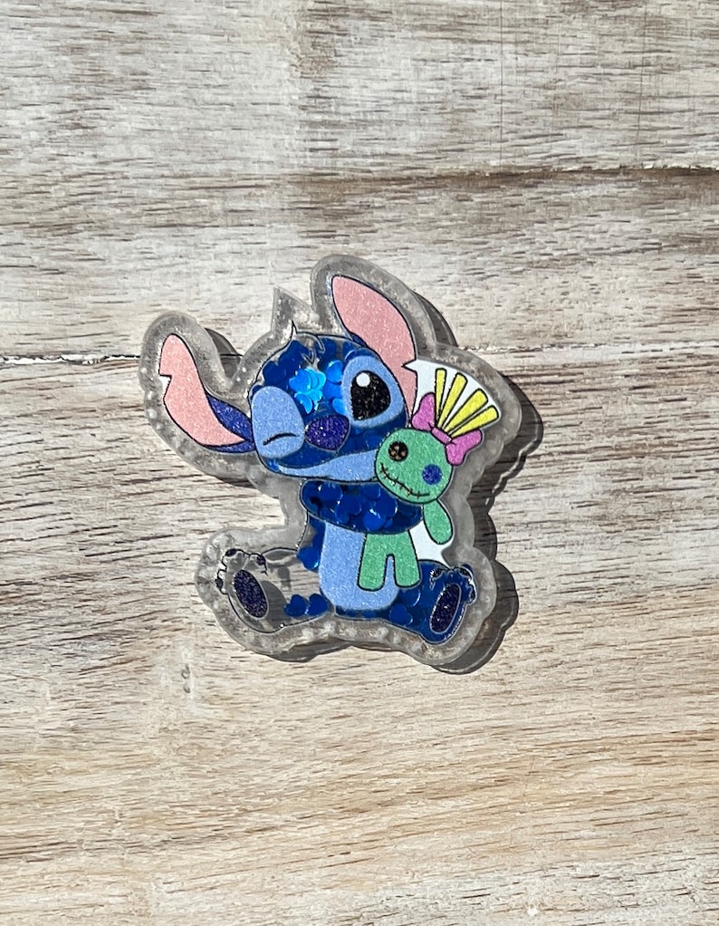 May include: A clear resin charm featuring a blue alien character with large ears and a green doll. The charm has a glittery, sparkly appearance with pink, blue, and yellow accents. The character is holding a small green doll with a yellow bow.
