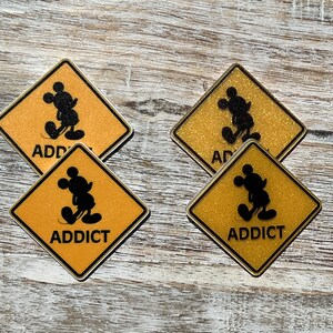 Puede incluir: Cuatro señales de advertencia amarillas en forma de diamante con bordes negros y una silueta negra de Mickey Mouse. Las señales tienen la palabra "ADDICT" impresa en letras negras.