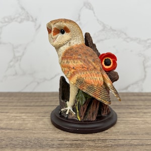 May include: A figurine of a barn owl perched on a branch with a red poppy flower. The owl is brown and white with orange markings. The figurine is on a brown base.