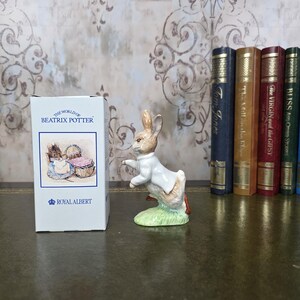 Peut inclure: Figurine en céramique de Pierre Lapin en veste blanche, sur une base verte. À côté, une boîte blanche avec l'inscription "THE WORLD OF BEATRIX POTTER" et "ROYAL ALBERT". Des livres en arrière-plan.
