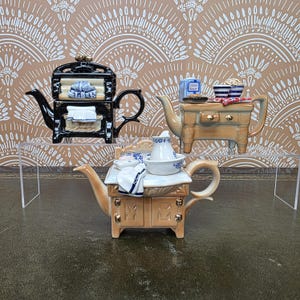 May include: Three ceramic teapots shaped like vintage kitchen stoves. One is black with white accents, while two are tan with gold-toned hardware. Each teapot features miniature kitchen items on top, including bowls, cloths, and a blue box labeled Caster Sugar.