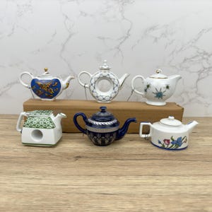 Puede incluir: Seis teteras de porcelana en miniatura en varios colores y dise&ntilde;os. Las teteras est&aacute;n dispuestas en una fila sobre una superficie de madera.