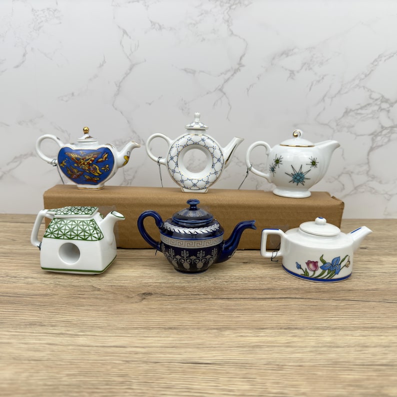 Puede incluir: Seis teteras de porcelana en miniatura en varios colores y dise&ntilde;os. Las teteras est&aacute;n dispuestas en una fila sobre una superficie de madera.