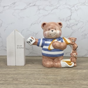 Puede incluir: Una tetera de cerámica con forma de oso de peluche con una camisa a rayas azules y blancas, sosteniendo una taza que dice "tea". La tetera tiene un asa con dos figuras de osos más pequeños. La tetera mide aproximadamente 15 cm de alto.