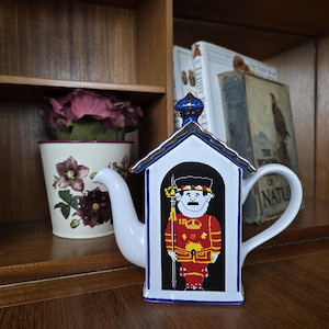 Peut inclure: Théière en céramique blanche en forme de maison, avec une illustration de garde royal en tenue rouge et or. La théière a un toit et une poignée bleus et dorés, et un long bec verseur.