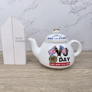 Könnte beinhalten: Weiße Keramik-Teekanne mit goldenen Verzierungen, mit dem Text "THE YEAR OF WAR AND PEACE" und "50 DAY". Enthält britische und amerikanische Flaggen sowie den Text "1945 AUG 15TH 1995". Die Teekanne ist etwa 15 cm hoch.