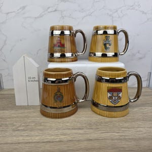 Puede incluir: Cuatro jarras de cerveza de cerámica con detalles y asas plateadas. Cada jarra presenta un escudo o emblema diferente, con texto como "University of Manchester". Las jarras miden aproximadamente 15 cm de alto.