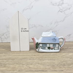 May include: A ceramic teapot shaped like a house, with a blue roof, white walls, and pink accents. The teapot is approximately 6 inches in height. A white, house-shaped object stands next to the teapot.