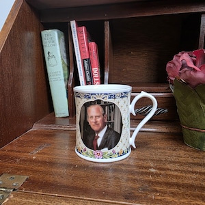 May include: A white ceramic mug featuring a portrait of a man in a suit, set within an ornate frame. The mug has a curved handle and is decorated with floral and geometric patterns. Books are visible in the background, suggesting a vintage or collectible theme.