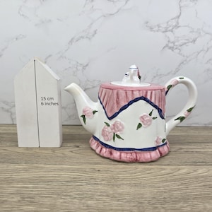 May include: A white ceramic teapot with a pink and blue floral design. The teapot features a pink lid and handle, with pink roses and green leaves. A white wooden house shape is next to the teapot, with the text "15 cm 6 inches".