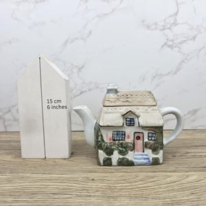 Peut inclure: Une théière en céramique en forme de cottage, avec un toit blanc, des fenêtres et une porte rose. La théière est décorée de vignes vertes et mesure environ 15 cm de haut. Un objet blanc en forme de maison se trouve à côté de la théière.