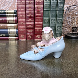Peut inclure: Une figurine en céramique représentant une petite souris brune portant un bonnet à carreaux roses et blancs, nichée dans une chaussure à talons bleu clair. La chaussure présente des accents décoratifs en forme de feuilles brunes sur le dessus.