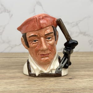 May include: A ceramic character mug depicting a man with a red hat and a tool in his hand. The mug is hand-painted and has a detailed design.