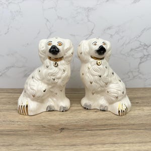 Puede incluir: Dos figuritas de perro de cerámica blanca con detalles dorados y ojos negros. Los perros están sentados con la cabeza girada hacia un lado.