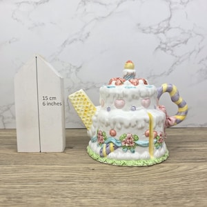 以下が含まれることがあります： 白いテクスチャード加工の釉薬が施された、ケーキ型の風変わりなティーポット。ピンクのハート、花の装飾、ろうそくが特徴です。ハンドルは黄色、紫、ラベンダーのストライプです。ティーポットの高さは約15cmです。