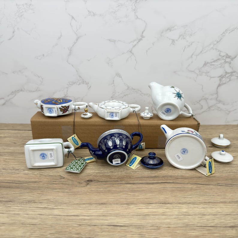 Puede incluir: Una colecci&oacute;n de seis teteras de porcelana en miniatura con varios dise&ntilde;os azules y blancos. Las teteras est&aacute;n dispuestas sobre una superficie de madera con sus fondos mirando hacia la c&aacute;mara. Cada tetera tiene una peque&ntilde;a etiqueta adjunta.