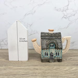 Op de afbeelding: Een decoratieve theepot in de vorm van een huisje, met een rieten dak en bakstenen accenten. De theepot is geschilderd in tinten blauw, groen en bruin. De hoogte van de witte doos is 15 cm (6 inch).