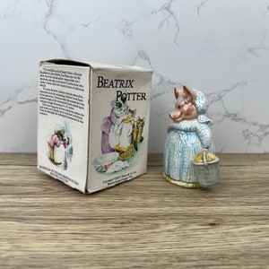 Może przedstawiać: Figurkę Beatrix Potter przedstawiającą świnię w niebiesko-białej sukience i białym czepeczku. Świnia trzyma wiaderko z żółtymi ziarnami. Figurka stoi na drewnianej powierzchni obok pudełka z logo Beatrix Potter i ilustracją kota i dwóch kociąt.