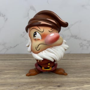 May include: A grumpy-looking dwarf figurine with a brown hat, a white beard, and a red and gold outfit. He has a large, pink nose and one eye is closed.