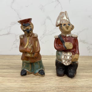 May include: Two ceramic figurines of soldiers. One soldier is wearing a red and brown uniform with a yellow sash and a black hat. The other soldier is wearing a red and white uniform with a white hat. Both figurines have painted details and are standing on a wooden surface.