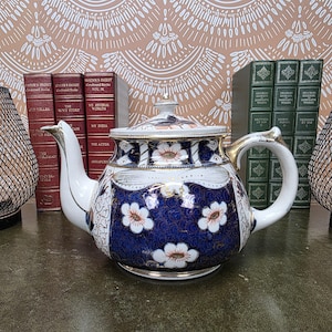 Puede incluir: Una tetera de porcelana con un patrón floral en azul oscuro y blanco con detalles en dorado. La tetera tiene un cuerpo redondeado, un pico curvo, un asa con detalles dorados y una tapa a juego con un pequeño pomo en la parte superior.