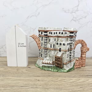 Könnte beinhalten: Eine Keramik-Teekanne in Form eines Schlosses, mit grauen und weißen Mauern, braunen Akzenten und einem grünen Sockel. Die Teekanne ist etwa 15 cm hoch. Der Griff ist gewölbt und der Ausguss detailliert.