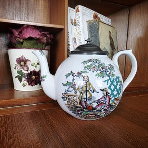 Könnte beinhalten: Weiße Keramik-Teekanne mit einer detaillierten, farbenfrohen Illustration von Figuren in einer Gartenlandschaft. Die Teekanne hat einen silbernen Deckel und einen gebogenen Griff. Eine Blumenvase und Bücher sind im Hintergrund.