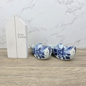 Può includere: Due zoccoli in ceramica con un design floreale blu su una superficie di legno. Gli zoccoli sono bianchi con motivi floreali blu. Un oggetto bianco a forma di casa, alto 15 cm, è a sinistra degli zoccoli.