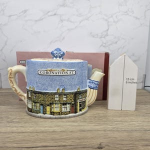 Könnte beinhalten: Keramik-Teekanne mit blauem Himmel-Design und der Aufschrift "CORONATION ST.". Die Teekanne zeigt detaillierte Gebäudeillustrationen und ist etwa 15 cm hoch. Griff und Ausguss sind verziert.