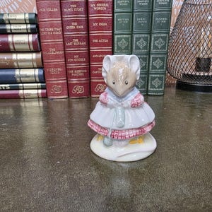 Puede incluir: Una figura de ratón de cerámica que lleva un vestido blanco con un patrón de cuadros rosas y un chal a juego. El ratón sostiene un proyecto de tejido verde con agujas. La figura está colocada frente a una colección de libros antiguos.