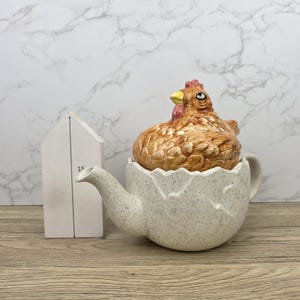 May include: A ceramic teapot shaped like a cracked eggshell, with a brown and tan chicken lid. The teapot has a speckled, off-white finish. A small, white, house-shaped object is to the left of the teapot.
