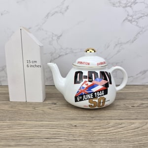 Könnte beinhalten: Weiße Keramik-Teekanne mit goldenem Griff und Deckelknauf. Die Teekanne zeigt ein Design zum 50. Jahrestag des D-Day mit Flaggen und Text. Der Text enthält "Operation Overlord" und "6. Juni 1944".