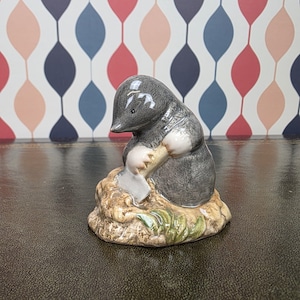 May include: A ceramic figurine of a mole digging with a shovel. The mole is gray and white, with a detailed face and paws. It is posed on a small mound of dirt and grass, with a colorful patterned background.